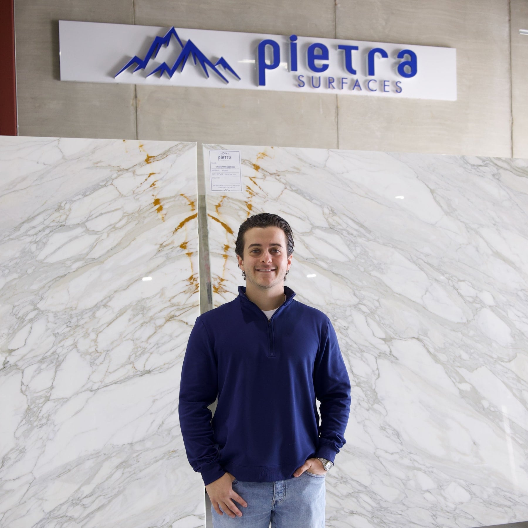 Standing in front of bookmatched marble slabs beneath the Pietra Surfaces sign